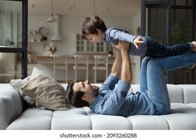 Happy young father lying on couch at home, lifting excited little child son, smiling excited dad and adorable kid boy having fun, practicing acroyoga in living room, engaged in funny activity