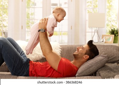 Happy Young Father Lying On Sofa, Lifting Baby Girl High Up, Smiling, Having Fun. Side View.