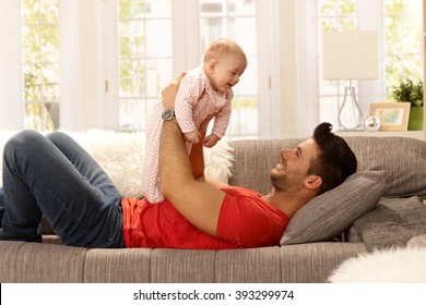 Happy Young Father Lying On Sofa At Home, Lifting Baby Daughter Up, Having Fun. Side View.