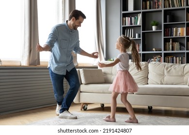Happy young father showing inviting dancing moves to smiling joyful little preschool kid daughter, preparing disco performance for kindergarten at home, hobby activity family pastime in living room.