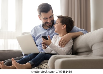 Happy Young Father Sit On Couch Using Laptop Relax With Preschooler Son Holding Smartphone Have Fun Together, Smiling Dad And Little Boy Child Enjoy Weekend At Home Rest On Sofa Busy With Gadgets