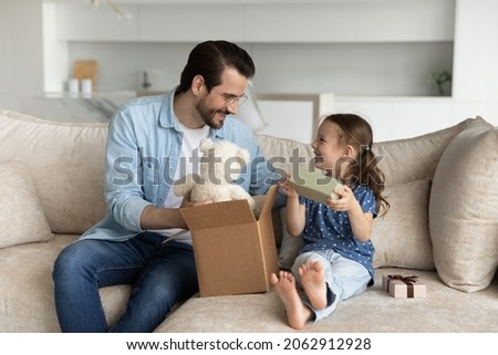 Happy young father unboxing parcel with gifts for small cute kid daughter, sitting together on cozy sofa in living room, satisfied with fast delivery purchase shipment or high quality fluffy toy.