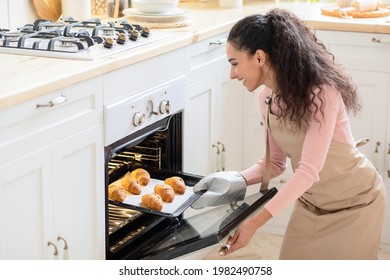 Happy Young Female Baking Croissants For Breakfast At Home, Smiling Millennial Woman Wearing Mitten And Apron Taking Tray With Fresh Baked Pastry Out Of Oven, Enjoying Cooking In Kitchen