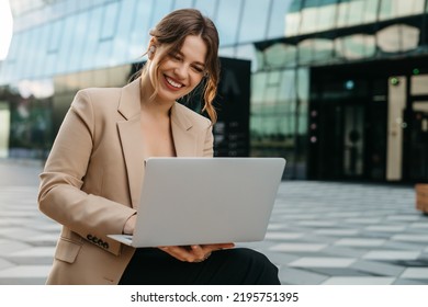 Happy young female blonde female student typing on laptop, talking on video call while sitting outdoors in city, business woman or female freelancer, working outdoors, cityscape in the background. - Powered by Shutterstock