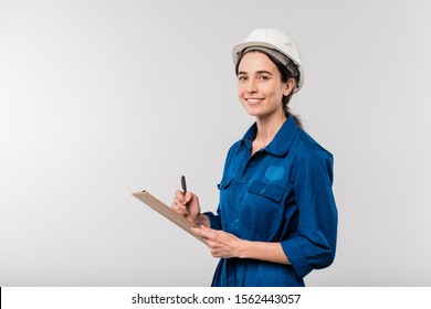 Happy Young Female Builder In Blue Workwear And Hardhat Making Notes In Document While Standing In Isolation