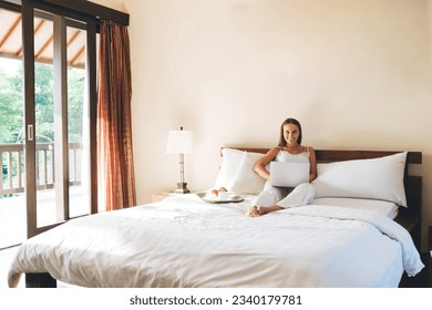 Happy young female in casual clothes smiling and looking at camera while using laptop on bed in bedroom at home - Shutterstock ID 2340179781