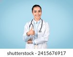 Happy young female doctor with stethoscope around her neck giving thumbs up and smiling, signifying satisfaction and positivity in the healthcare environment, blue backdrop