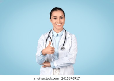 Happy young female doctor with stethoscope around her neck giving thumbs up and smiling, signifying satisfaction and positivity in the healthcare environment, blue backdrop - Powered by Shutterstock