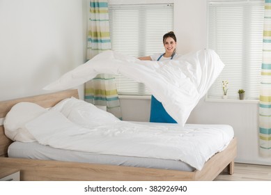 Happy Young Female Housekeeper Changing Bedsheet On Bed In Room