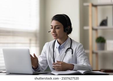 Happy Young Female Indian Family Doctor Therapist In Headphones With Mic Consulting Patient Online By Computer Video Call Conversation, Telling Advices Or Taking Part In Educational Virtual Event.