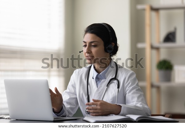 Happy young female Indian family doctor therapist\
in headphones with mic consulting patient online by computer video\
call conversation, telling advices or taking part in educational\
virtual event.