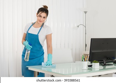 Happy Young Female Janitor Using Spray To Wipe Desk