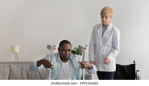 Happy Young Female Nurse Provide Care, Medical Service, Help And Support. Serious Adult Lady Looks At African American Mature Man With Crutches Get Up From Couch, Near Wheelchair At Home, Panorama