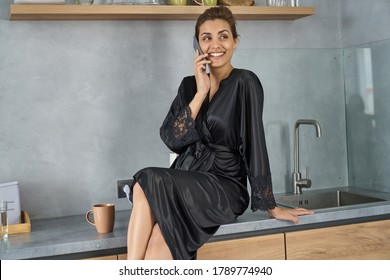 Happy Young Female In Silk Black Dressing Gown Is Sitting On Table In Kitchen And Communicating On Cell Phone