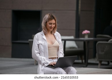 Happy young female student sits on a bench and watches a webinar, attends an online class, study conference, talks with a teacher via video link, laughs, writes, studies on a city street - Shutterstock ID 2165510553