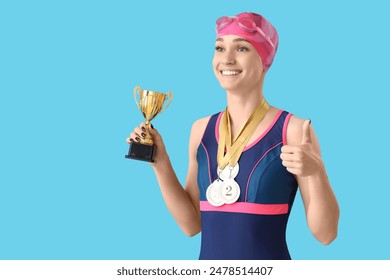 Happy young female swimmer with medals and gold cup showing thumb-up on blue background - Powered by Shutterstock