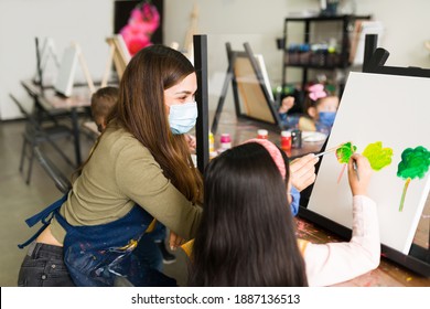 Happy young female teacher and an elementary girl with face masks are painting a landscape of trees on a canvas during an art class - Powered by Shutterstock