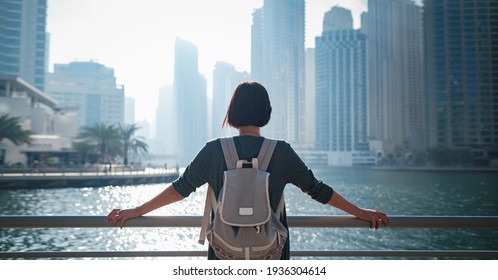 Happy young female traveler in the big city of Dubai, famous place Dubai marina. Luxury and comfortable tourism season in United Arab Emirates. Back or rear view of young woman in dress and backpack