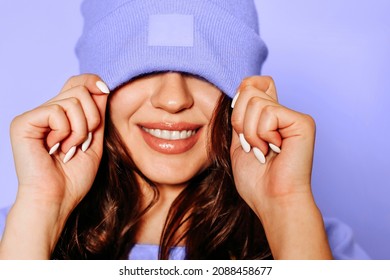 Happy young female in trendy yellow sweatshirt and knitted hat covering eyes smiling brightly against very peri background