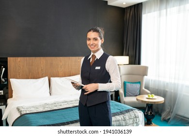 Happy Young Female Worker Of Luxurious Hotel In Uniform Standing In Front Of Camera Against Bed And Using Touchpad While Looking At You