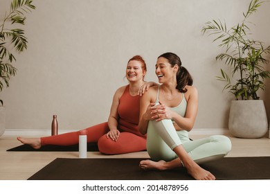 Happy young females sitting on gym floor and smiling. Two women taking a break from workout at fitness studio.