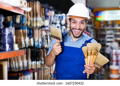 Happy Young Foreman In Blue Overalls Choosing Brushes In Paint Store