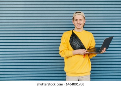 Happy Young Freelancer Stands With An Open Laptop In His Hands Against The Background Of A Blue Wall, Looks At The Camera And Smiles, Wears Casual Clothes, Cap And Fanny Pack. Copyspace