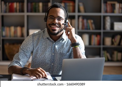 Happy young friendly african american man in eyeglasses wearing headset with mic, looking at camera. Smiling professional financial advisor consulting clients online, solving problems issues.