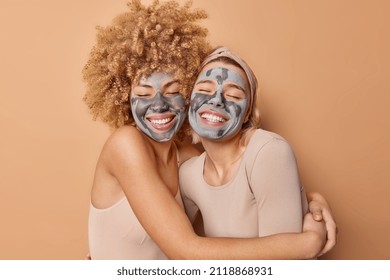 Happy young friendly women embrace each other with love smile broadly apply moisturizing clay facial masks for skin treatment dressed in casual clothes isolated on brown background. Face care concept