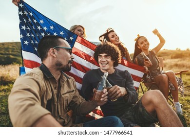 Happy young friends are celebrating Fourth of July and enjoy a sunny day at the mountain. They're holding American flag, laughing and toasting with beer bottles. - Powered by Shutterstock