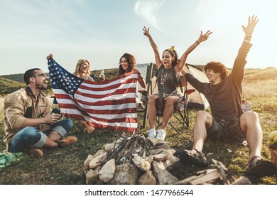 Happy young friends are celebrating Fourth of July and enjoy a sunny day at the mountain. They're holding American flag, laughing and toasting with beer bottles. - Powered by Shutterstock