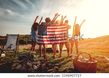 Happy young friends enjoy a sunny day in nature. They're looking at sun holding american flag and greeting, happy to be together.