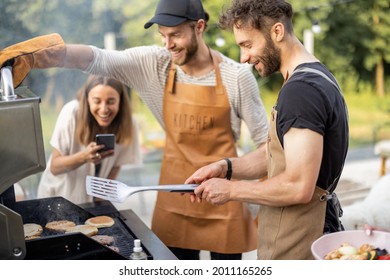 Happy Young Friends Hanging Out Together, Grilling Meat And Buns For Burgers On A Modern Grill At Picnic. People Cooking Food Outdoors