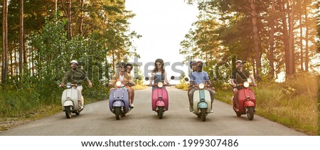 Happy young friends riding scooter motorbikes in row along asphalt road in sunny nature, panorama. Leisure, fun and entertainment concept