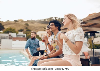 Happy Young Friends Sitting By The Pool Having Cocktail. Young People Relaxing By The Swimming Pool With Their Feet In Water During A Party. Men And Women Enjoying Rooftop Party.