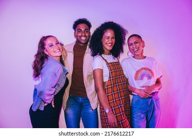 Happy Young Friends Smiling At The Camera At A House Party. Group Of Multicultural Young People Standing Together In Bright Neon Light. Vibrant Friends Having A Good Time On The Weekend.