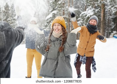 Happy Young Friends In Winter Coats Having Snowball Fight In Forest: Excited Girl Standing In Center And Feeling Snow On Face