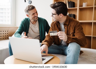 Happy Young Gay Couple Using Laptop Computer While Sitting On A Couch At Home, Shopping Online With A Credit Card