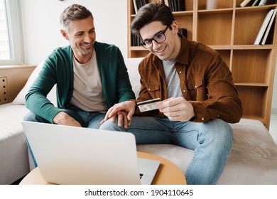 Happy young gay couple using laptop computer while sitting on a couch at home, shopping online with a credit card