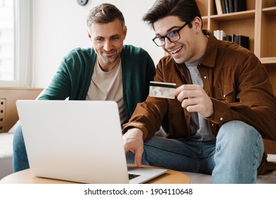 Happy Young Gay Couple Using Laptop Computer While Sitting On A Couch At Home, Shopping Online With A Credit Card