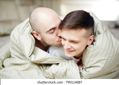 Happy Young Gay Men Wrapped In Blanket Lying In Bed While Spending Time Together