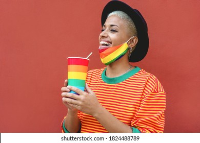 Happy young gay woman drinking from a rainbow glass while wearing a pride mask - Focus on girl face - Powered by Shutterstock