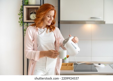 Happy, young, ginger girl in bright outfit standing and making her morning tea using electric kettle in the modern kitchen - Powered by Shutterstock