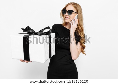 Happy young girl in a black dress and sunglasses, talking on the phone holding a gift with a black ribbon black Friday, on a white background