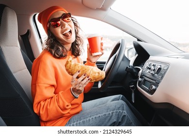 A Happy Young Girl Driver Is Drinking Coffee And Eating A Fresh Croissant While Driving A Car. The Concept Of Ordering Fast Food Delivery And Dangerous Driving