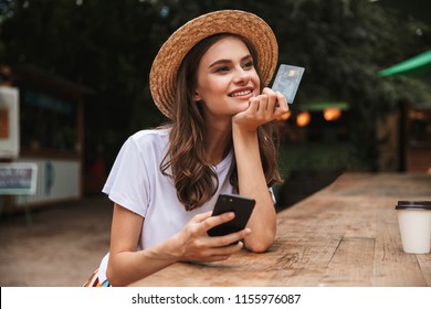 Happy Young Girl Holding Plastic Credit Card While Using Mobile Phone At The Cafe Outdoors