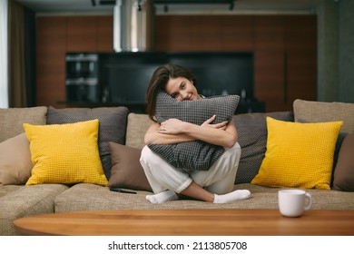Happy young girl hugging soft pillow sitting on comfortable sofa. Relaxed female resting in modern living room at home