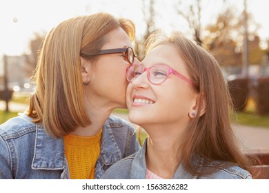 Happy young girl laughing as her mom kissing her on the cheek. Cheerful mother and daughter outdoors in autumn