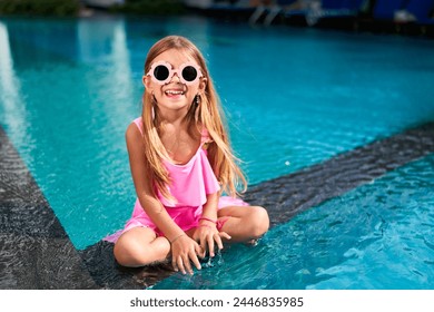 Happy young girl in pink swimsuit sits by pool edge, feet dipped in water, wearing oversized round sunglasses. Summer vibes, playful child enjoys poolside fun, relaxed sunny day at resort. - Powered by Shutterstock