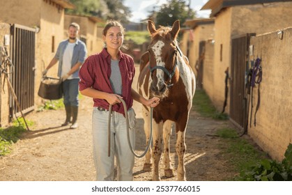 Happy young girl preparing horse for riding and walking in pasture on small farm - Powered by Shutterstock